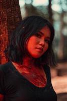 un asiatique femme dans noir vêtements enduit sa corps avec rouge du sang avant le rituel commence dans de face de une gros arbre photo