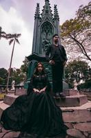 une couple de les amoureux séance et permanent dans de face de une funéraire monument dans tout noir vêtements et effrayant expressions sur Halloween photo