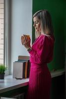 une belle jeune femme blonde en costume rose-rouge vif se tient près de la fenêtre de son bureau, se repose et boit du café chaud dans une tasse. photo