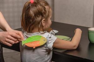 le plus âgée sœur colles une papier poisson à sa peu sœur sur sa retour à Accueil dans le cuisine, le enfant a petit déjeuner. marrant famille blagues, avril imbécile journée fête photo