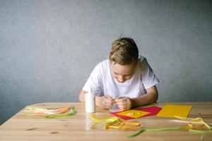 mignonne garçon fait du fleurs pour une carte postale par quilling tandis que séance à une table photo