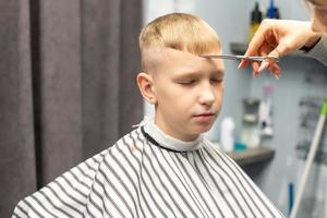 une écolier est séance dans une salon de coiffure, Faire le sien cheveux avec les ciseaux pour coupes de cheveux photo