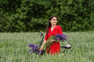 une jolie fille dans une rouge robe des stands avec une bouquet de lupins dans une champ suivant à une vélo photo