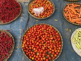 divers des légumes tomates, Chili, rouge oignon, maïs, carotte, chaux, Ail étant vendu à asiatique traditionnel marché. coloré des légumes sur rond bambou plateau à traditionnel marché sol photo