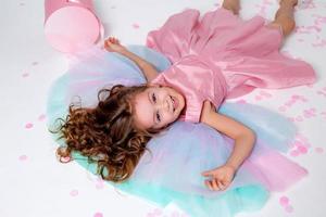 magnifique peu fille dans une élégant rose robe mensonges sur le sol jonché avec confettis. Haut voir. mode et style. enfant fête le sien anniversaire. photo dans le studio. espace pour texte. haute qualité photo