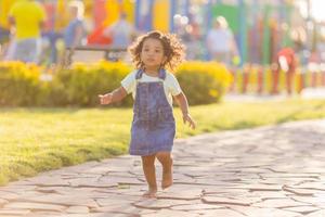 portrait peu content espiègle bambin basané fille dans une denim robe d'été permanent dans le jardin sur une ensoleillé journée. en marchant dans le Frais air. concept de une content enfance. espace pour texte. haute qualité photo