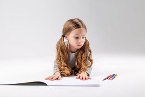 peu fille dans une rayé robe dessine dans un album mensonge sur le sol. enfant développement. mode de vie. préscolaire et école éducation. espace pour texte. haute qualité photo