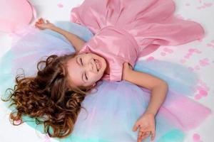 magnifique peu fille dans une élégant rose robe mensonges sur le sol jonché avec confettis. Haut voir. mode et style. enfant fête le sien anniversaire. photo dans le studio. espace pour texte. haute qualité photo