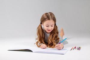 peu fille dans une rayé robe dessine dans un album mensonge sur le sol. enfant développement. mode de vie. préscolaire et école éducation. espace pour texte. haute qualité photo