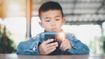 asiatique garçon à la recherche à le mobile téléphone écran attentivement. hyperstimulé les enfants concept. aussi beaucoup écran temps. 5 ans garçon en train de regarder vidéos tandis que la télé est travail.smartphone, l'Internet dépendance concept. photo