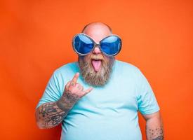 graisse homme avec barbe, tatouages et des lunettes de soleil fait du le geste de le cornes photo