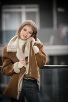 magnifique souriant fille dans élégant vêtements posant sur moderne balcon avec incroyable vue sur ville .portrait de une élégant à la mode blond adolescent avec fourrure manteau avec grand colliers portant dans balcon photo