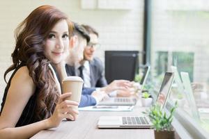Businesswoman buvant du café en regardant son ordinateur portable pendant que les collègues interagissent en arrière-plan photo