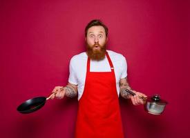 étonné chef avec barbe et rouge tablier cuisiniers avec la poêle et pot photo