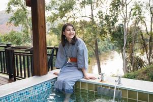 Jeune asiatique femme dans peignoir de bain séance sur Naturel chaud printemps bassin parmi le la nature à le recours photo
