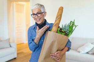 content mature femme en portant une papier sac plein de les courses de le supermarché. personnes âgées femme avec les courses à Accueil photo