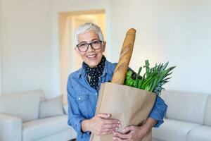 en bonne santé positif content femme en portant une papier achats sac plein de fruit et des légumes. portrait de magnifique Sénior femme épicerie achats sac avec des légumes à Accueil photo