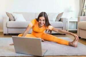 fille formation à maison, Faire faire des exercices et en train de regarder vidéos sur ordinateur portable, formation dans vivant chambre. magnifique femme est étirement sa jambes. elle est en train de regarder vidéo sur le l'Internet et répéter le Tâches photo