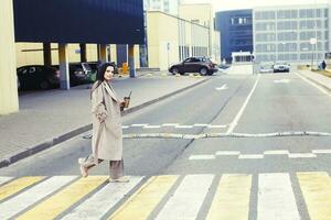 femme en portant café et traversée le route à piéton traversée contre Urbain ville Contexte photo