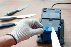 aiguiser un couteau sur un aiguiseur électrique à la maison. la main de l'homme enfonce la lame du couteau entre les aiguiseurs bleus, la poussière vole sur la machine. photo