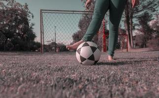 jambes de femme dans bleu pantalon, pieds nus dans milieu de parc en jouant avec une football Balle avec une football objectif avec filets et des arbres dans Contexte pendant le Matin photo