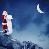 Père Noël claus lit le liste de cadeaux sur le de pointe de une Montagne en dessous de le lune photo