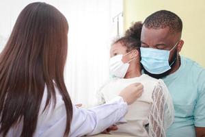 asiatique femelle médecin les usages une stéthoscope vérifier le battement de coeur de une peu Afro-américain fille séance avec sa père. concept de traitement pour infectieux maladies dans enfants, pendant coronavirus photo