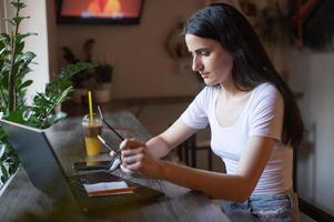 le fille est séance dans une café à le bar, à la recherche à le téléphone, en portant des lunettes dans sa main, une ordinateur portable photo