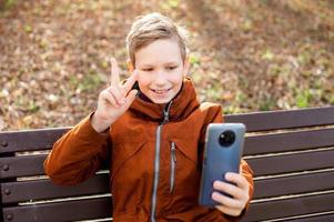 le garçon est parlant par vidéo sur le téléphone, séance sur une banc et en riant photo