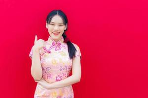 magnifique asiatique femme porte rose cheongsam et bras franchi tandis que regards à à caméra et sourit Heureusement et agissant main spectacles les pouces en haut avec le rouge fond, célébrer chinois Nouveau année thème. photo