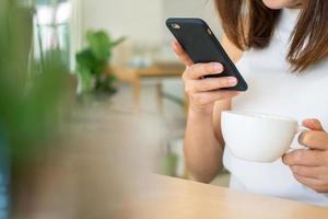 une femme est séance et en buvant café dans le maison et en utilisant une mobile téléphone à analyse pour un en ligne magasin. achats. achat des idées en ligne est vite et pratique. non avoir besoin à aller à le magasin. photo