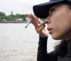 portrait femme fille adolescent Jeune Asie un la personne portant une chapeau et longue noir cheveux portant une noir chemise main tenir fumeur cigarette blanc Couleur permanent Extérieur par le mur Extérieur photo
