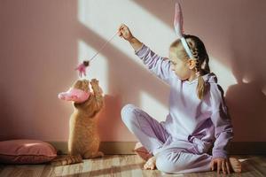 une écolière sur la tête un bandeau avec des oreilles de lapin joue avec un chat domestique rouge dans un chapeau de pâques au soleil photo