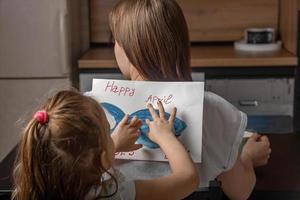 de bonne humeur peu fille collé une pièce de papier poisson à sa mère sur sa retour à Accueil dans le cuisine. marrant famille blagues, avril imbécile journée fête photo