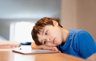 école enfant en utilisant tablette pour devoirs, enfant ennuyé visage mensonge tête vers le bas à la recherche en dehors Profond dans pensée, Jeune garçon apprentissage en ligne classe pièce à maison, e-learning ou homeschooling éducation concept photo