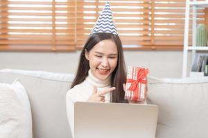 célébrer sur journée de content anniversaire saison, jolie asiatique Jeune femme en utilisant portable ordinateur, salutation sur vidéo appel pendant social distance, en portant cadeau boîte, content à avoir présent sur canapé, canapé à maison. photo