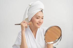 content beauté blogueur concept, mignonne asiatique Jeune femme, fille sourire, faire en haut visage par appliquant brosse rougir poudre sur sa joue, à la recherche à le miroir. gens Regardez avec Naturel mode style. photo