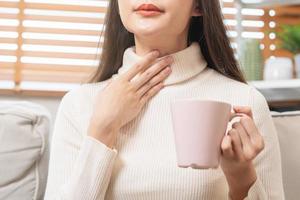 malade, mal asiatique Jeune femme, fille main toucher cou, avoir endolori gorge, en portant verre ou tasse, boisson chaud eau, avoir une fièvre, grippe dans faiblesse, séance sur canapé à maison. santé se soucier sur virus saisonnier. photo