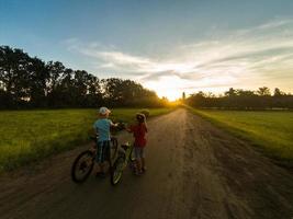 enfant équitation un bicyclette ensemble sur ensoleillé été soir. séance sur vélo étagère. famille de deux gens profiter en voyageant dans scénique champ plus de le coucher du soleil ciel Contexte. photo