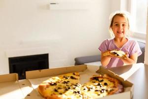 charmant content Jeune fille rire et mordant de gros tranche de Frais fabriqué Pizza. elle asseoir à blanc chaise dans Provence style intérieur, sourire et prendre plaisir ensoleillé journée et délicieux repas. elle a longue blond cheveux. photo