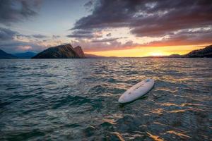 une planche de surf mensonges dans le l'eau dans une magnifique paysage tandis que le Soleil ensembles en quittant une rouge ciel derrière photo