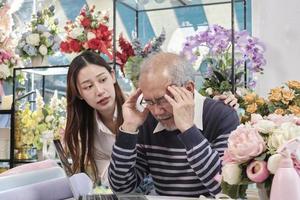 asiatique personnes âgées Masculin fleuriste propriétaire est indisposé avec maux de tête et santé se détériore, stress de surmenage, Jeune femelle ouvrier soutien avec se soucier dans une fleur boutique, famille pme affaires entrepreneur. photo