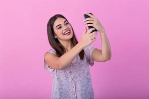 mignonne magnifique fille avec longue cheveux dans studio avec rose des murs fabrication une selfie. elle regards à sa téléphone intelligent et fait du une selfie. photo