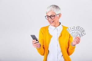 portrait de une Enchanté Sénior femme en portant bouquet de argent billets de banque et payant en ligne sur sa mobile téléphone plus de gris Contexte photo