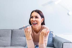 femme séance sur canapé détient téléphone pourparlers sur haut-parleur avec ami, fait du voix reconnaissance ou demande les usages l'Internet prestations de service par virtuel assistant, record l'audio message, traducteur app concept photo