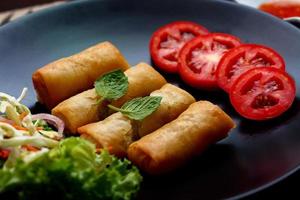 frit printemps Rouleaux avec des légumes et tomates mis dans une noir assiette sur une noir en bois table et plongement sauce. photo