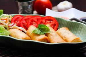 frit printemps Rouleaux, des légumes et tomates mis dans une vert feuille forme assiette sur une noir en bois table et plongement sauce. photo