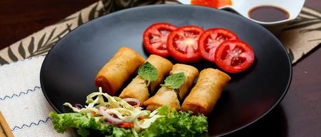 frit printemps Rouleaux avec des légumes et tomates mis dans une noir assiette sur une noir en bois table et plongement sauce. photo