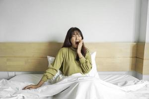 mignonne asiatique femme élongation en dehors et bâillement tandis que séance sur sa lit dans brillant chambre photo