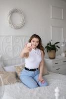 une mignonne fille est assis sur une blanc lit avec une téléphone et prend une selfie photo